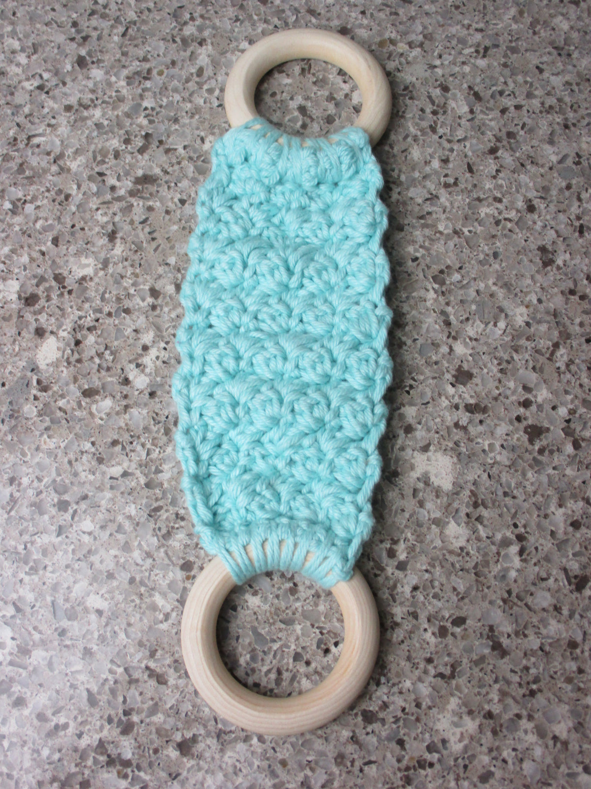 A handmade Kitchen towel Holder made of 100% Cotton Yarn in Med. Aqua Color with two cream-colored wooden rings, shown on a speckled gray countertop.