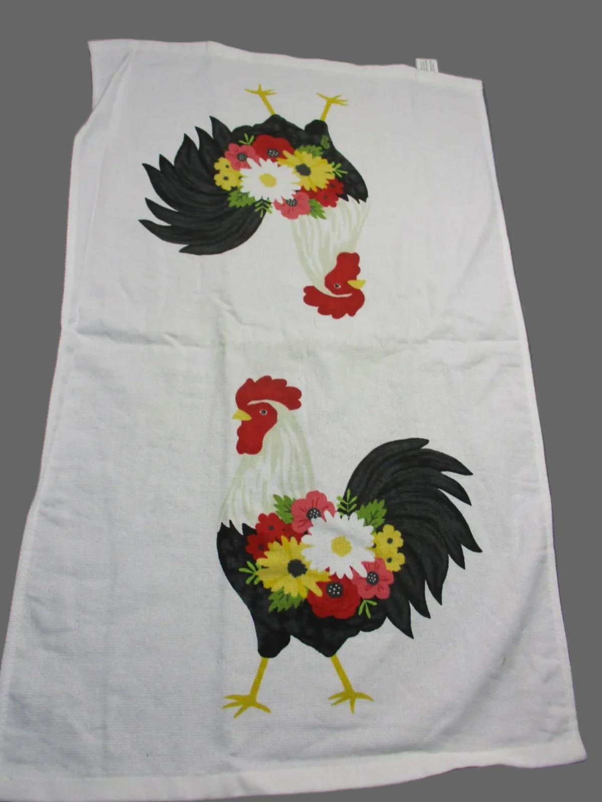 A white fabric towel laid fully opened on a gray surface, featuring two large black rooster designs with colorful flower bouquets in red, yellow, white, and green.
