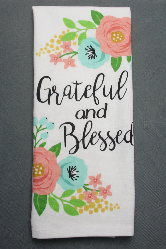 A folded white fabric item, likely a tea towel, printed with the words 'Grateful and Blessed' in black script, surrounded by a colorful floral design in pink, teal, yellow, and green, laid flat on a dark gray background.