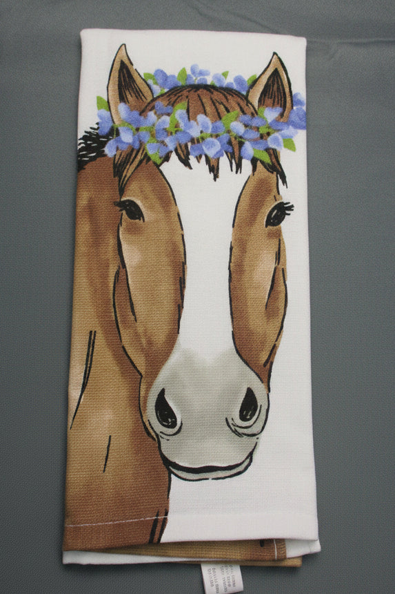 A folded white kitchen towel featuring a printed illustration of a brown horse with blue flower crowns, laid flat on a dark gray fabric background.