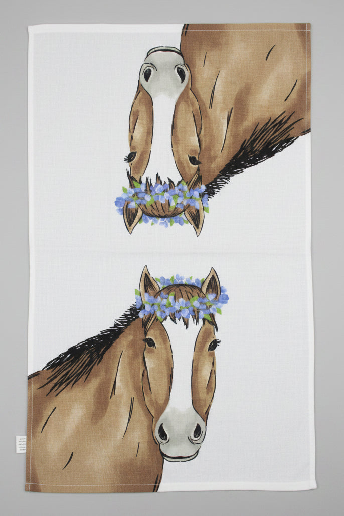 A white towel fully opened featuring a printed design of two brown horses facing each other, each wearing a blue flower crown, laid flat on a dark gray fabric surface.