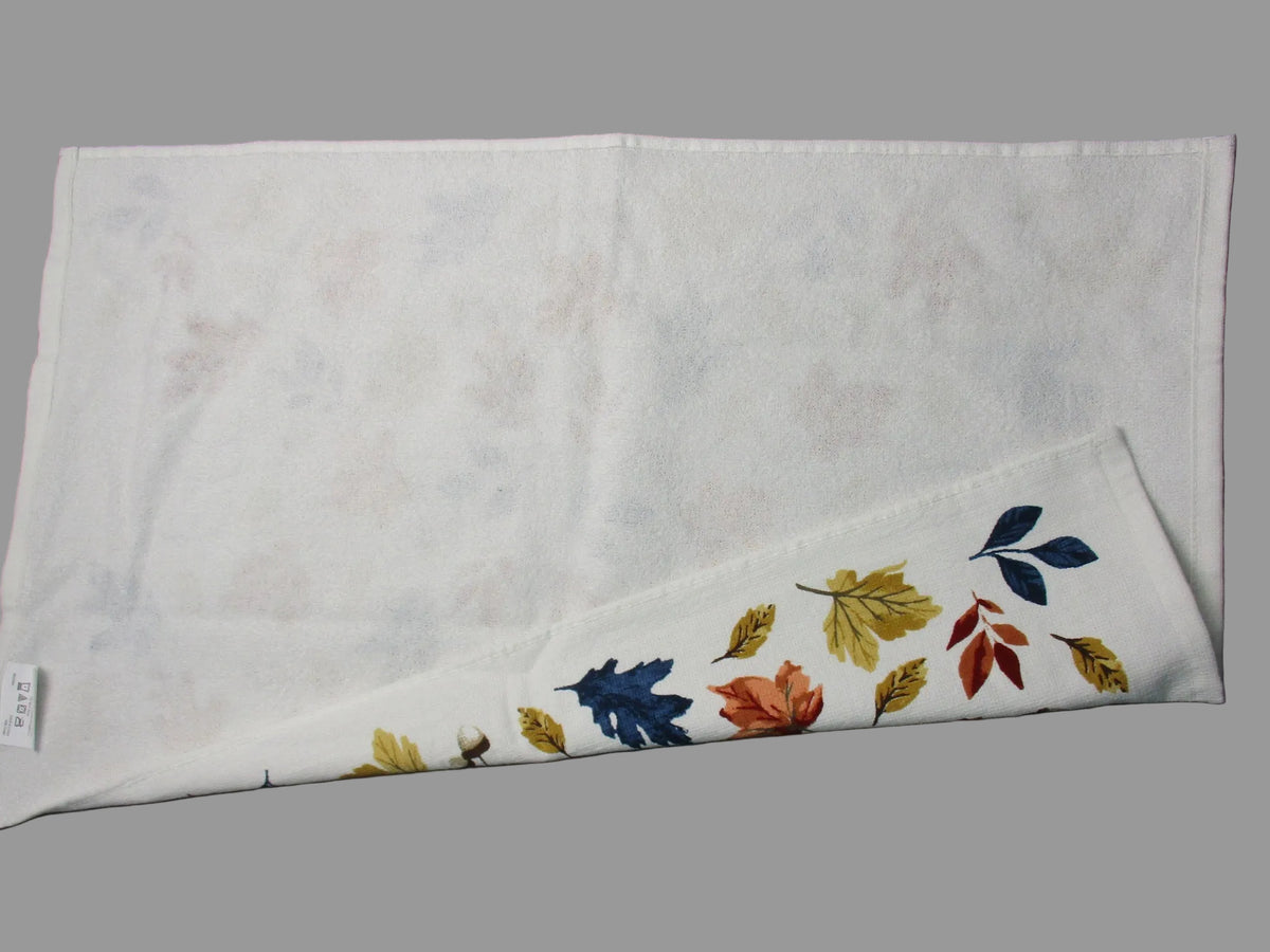 back side of white cotton tea towel with a subtle tonal floral pattern, folded to show a printed border of autumn leaves in blue, yellow, and orange, laid flat on a dark gray surface.