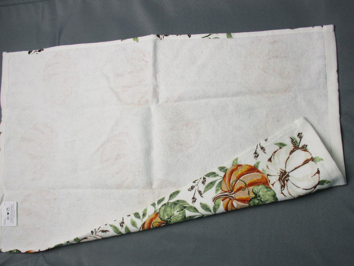 Back side of towel with a printed autumn pumpkin and leaf pattern, folded diagonally on a dark gray surface.