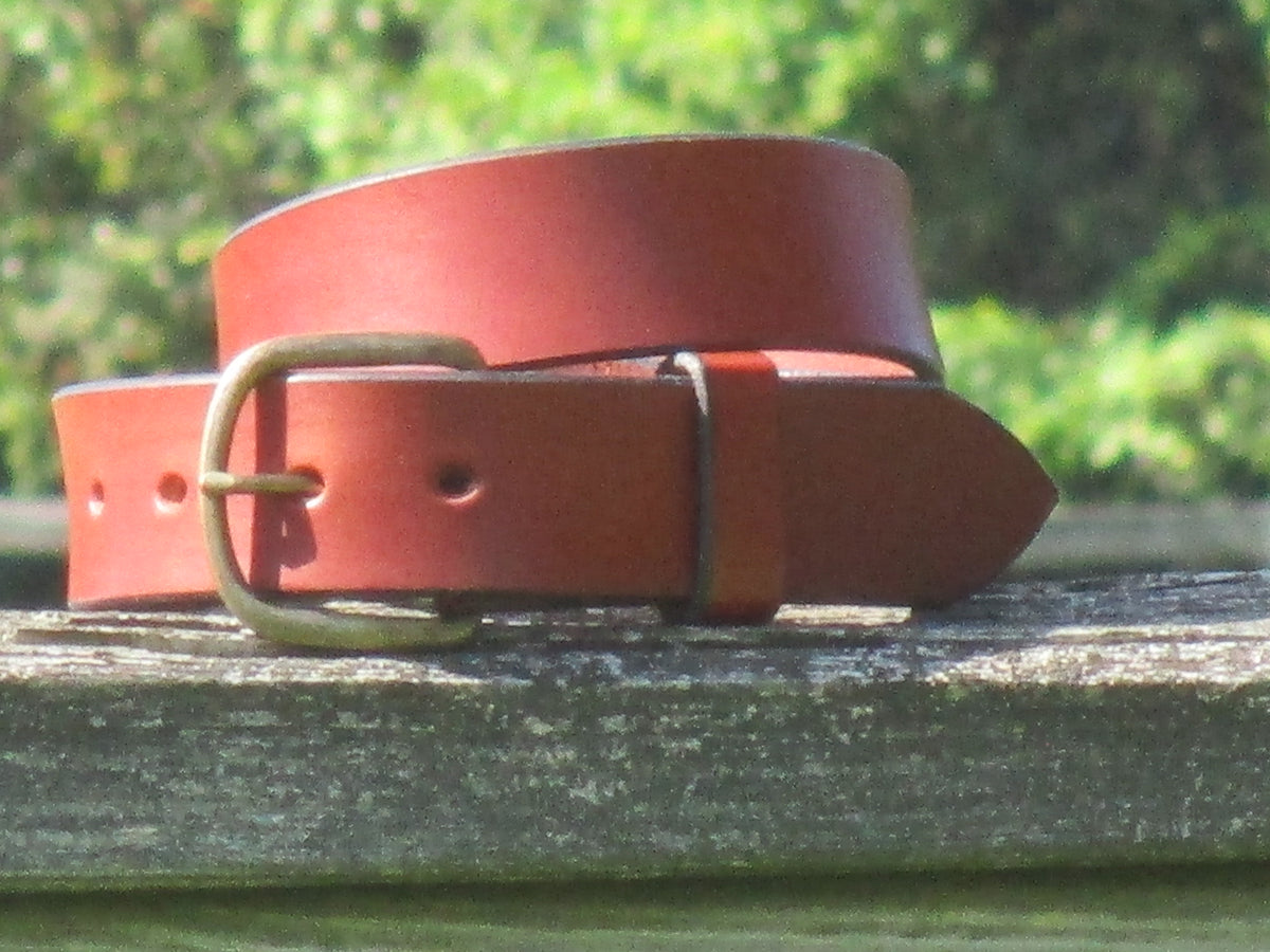 Chestnut leather belt made from 9/10 English Bridle Leather with an Antique Brass buckle on a wooden surface with a blurred green background