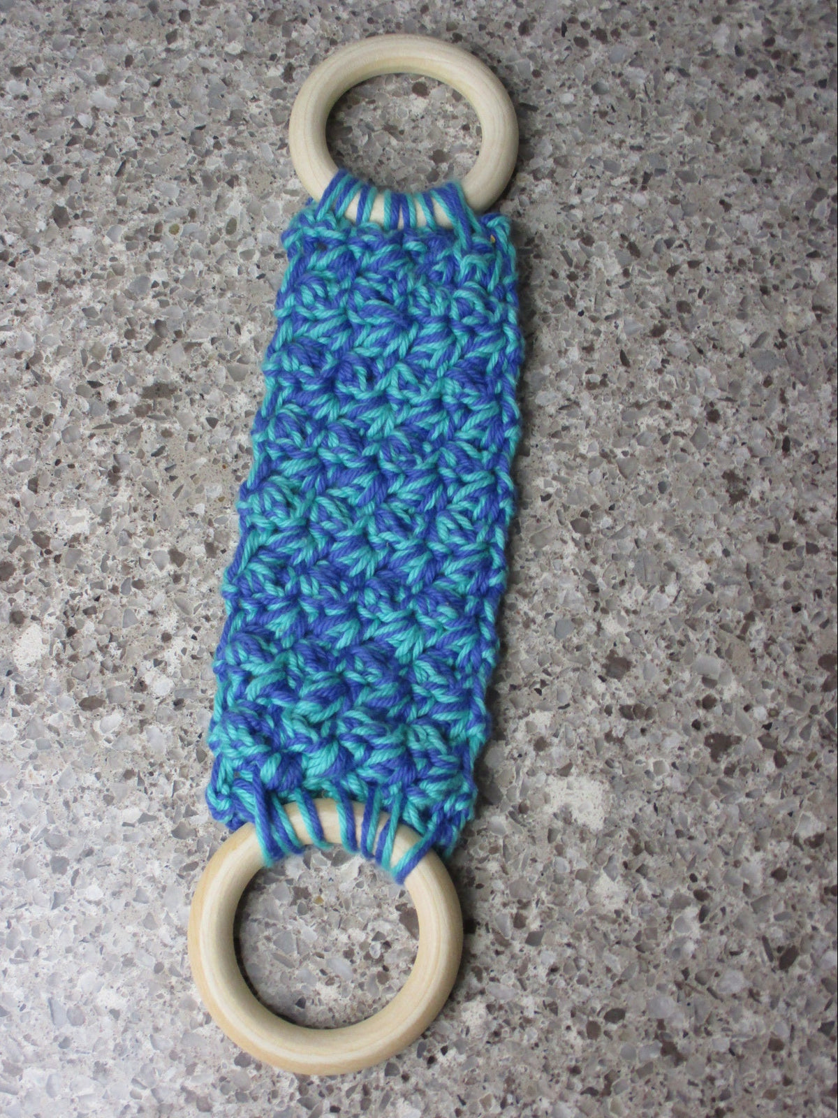 A handmade Kitchen towel Holder made of 100% Cotton Yarn in Cobalt Blue and Med. Teal Colors with two cream-colored wooden rings, shown on a speckled gray countertop.