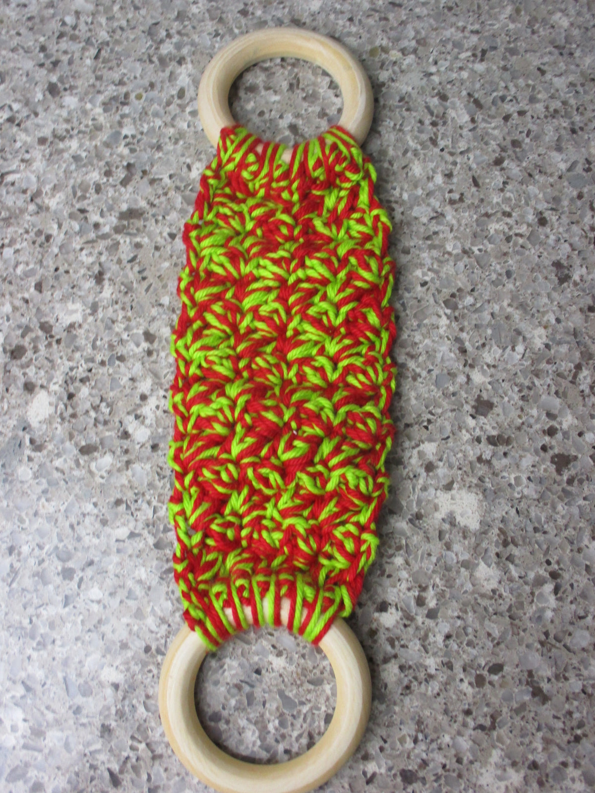 A handmade Kitchen towel Holder made of 100% Cotton Yarn in Bright Red and Lime Green Colors with two cream-colored wooden rings, shown on a speckled gray countertop.