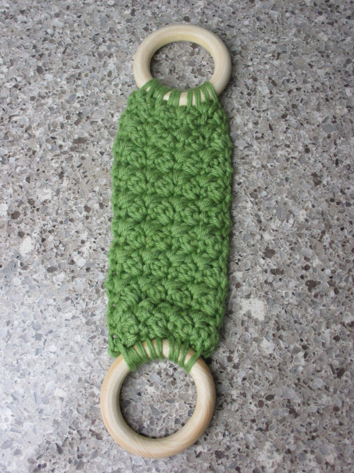 A handmade Kitchen towel Holder made of 100% Cotton Yarn in Olive  Green Color with two cream-colored wooden rings, shown on a speckled gray countertop.