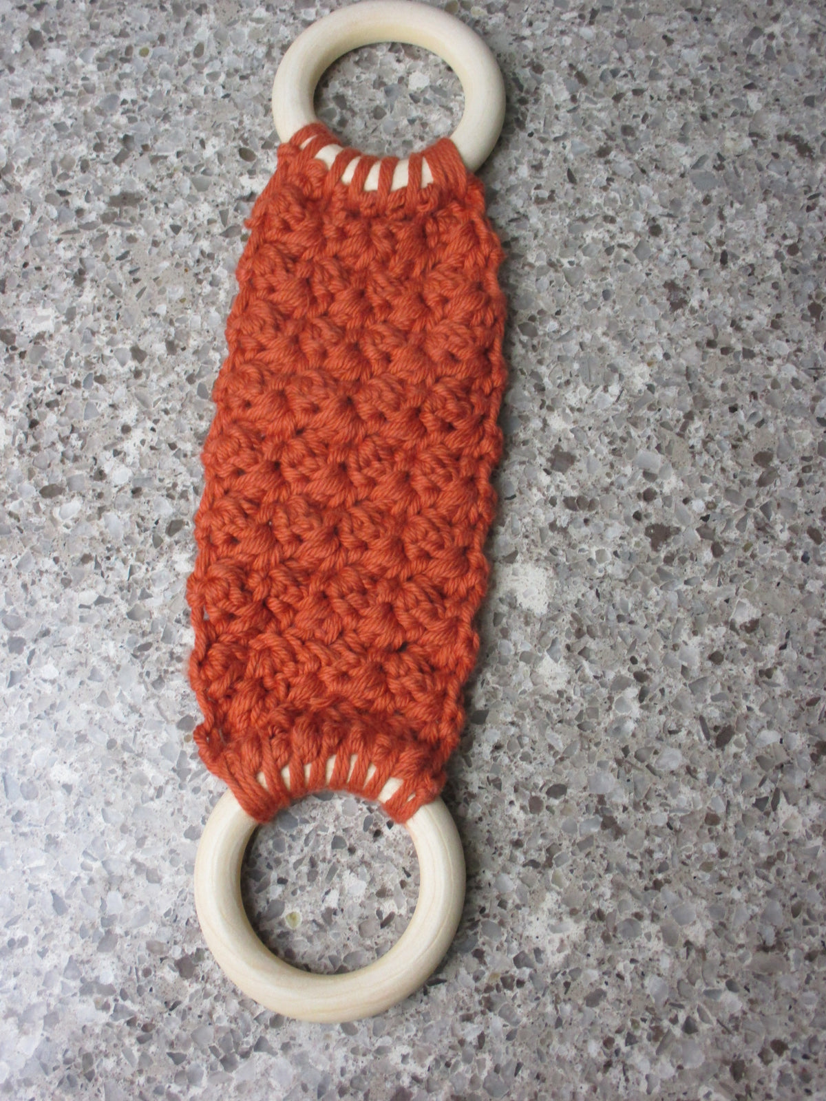 A handmade Kitchen towel Holder made of 100% Cotton Yarn in Rust Color with two cream-colored wooden rings, shown on a speckled gray countertop.
