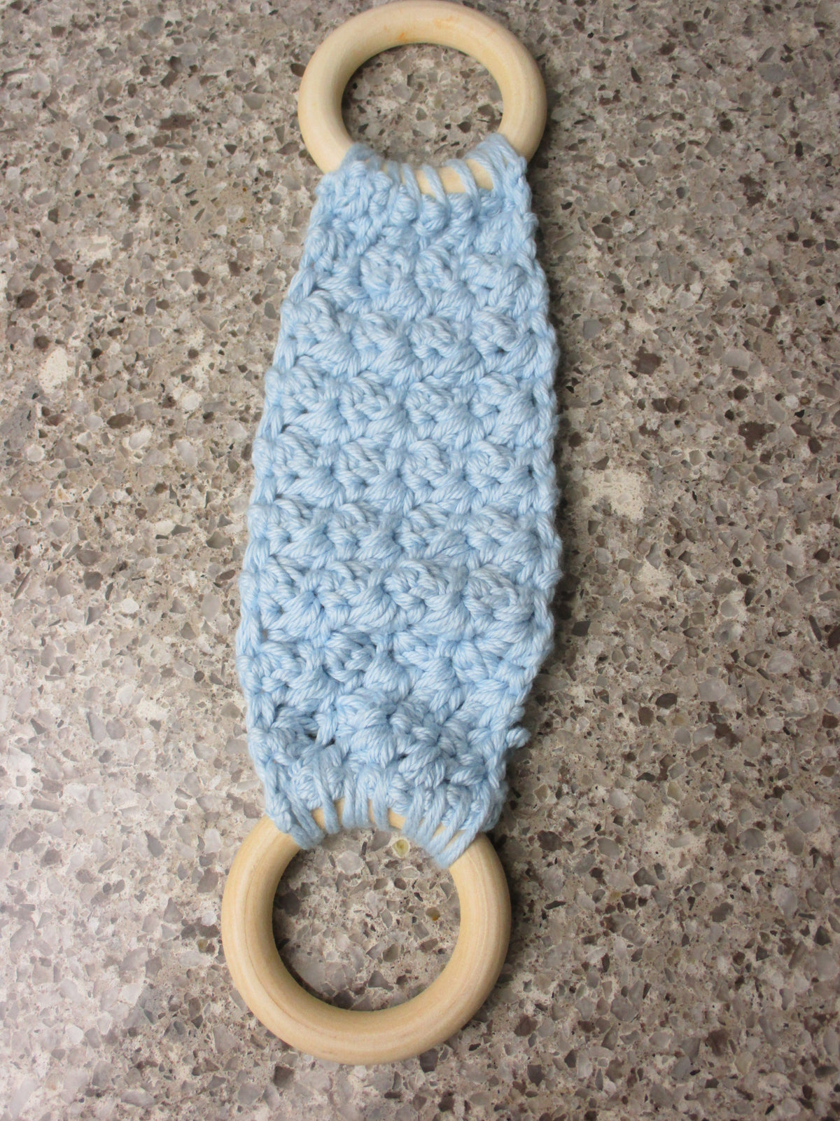 A handmade Kitchen towel Holder made of 100% Cotton Yarn in Soft Blue Color with two cream-colored wooden rings, shown on a speckled gray countertop.