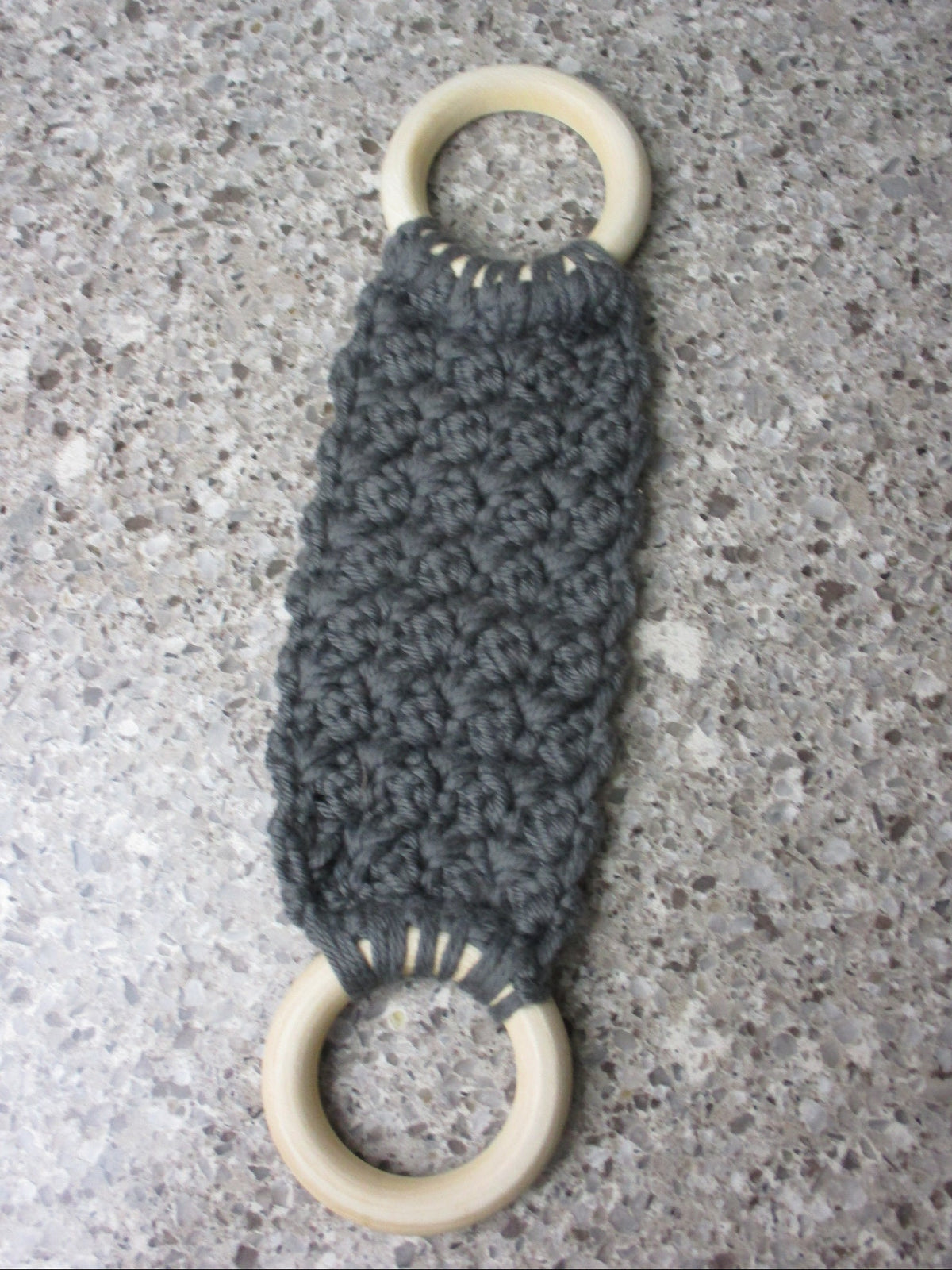 A handmade Kitchen towel Holder made of 100% Cotton Yarn in Dk. Gray Color with two cream-colored wooden rings, shown on a speckled gray countertop.