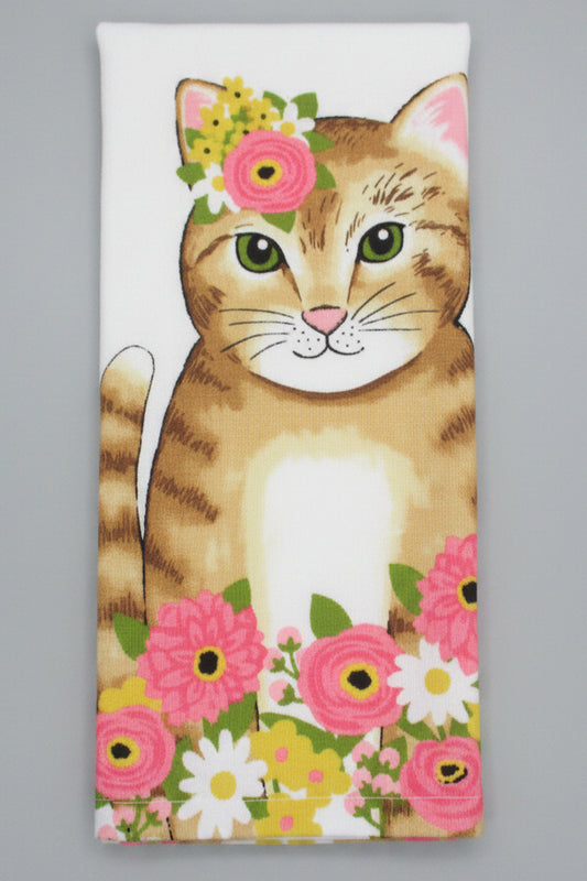 A folded white towel, featuring a printed design of a brown tabby cat with green eyes surrounded by pink, yellow, and white flowers, laid flat on a dark gray background.