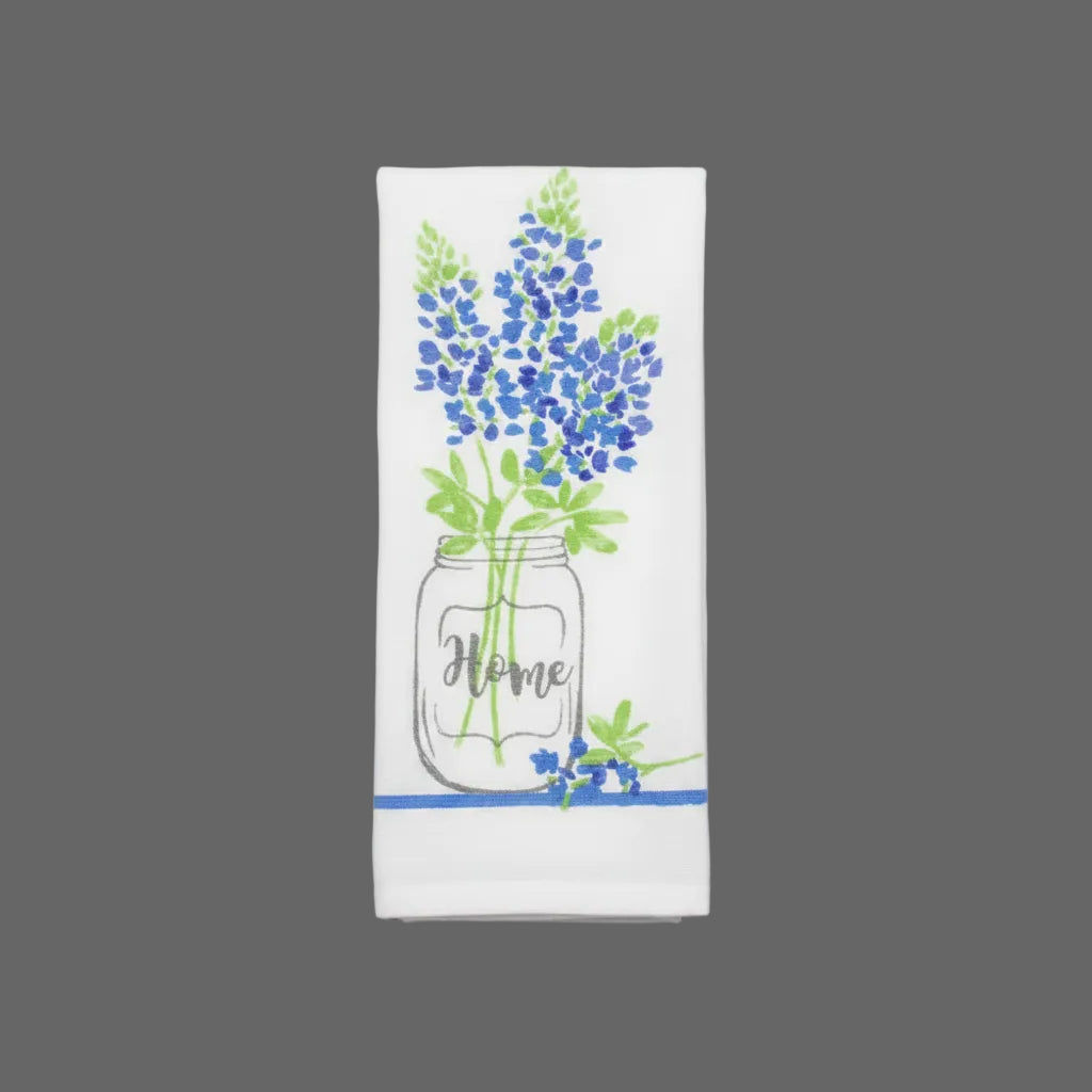 A folded white kitchen towel featuring a printed design of bluebonnets in a mason jar with the word 'Home' written inside, set against a dark gray fabric background.
