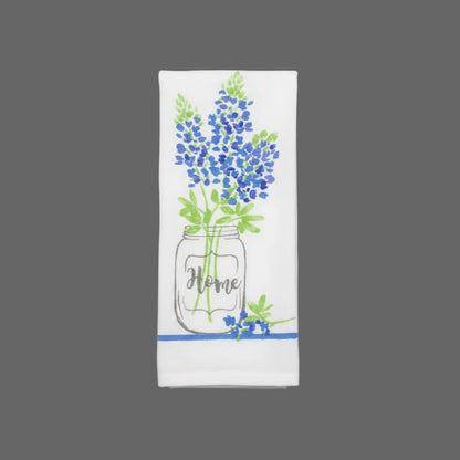 A folded white kitchen towel featuring a printed design of bluebonnets in a mason jar with the word 'Home' written inside, set against a dark gray fabric background.