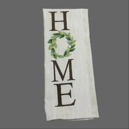 A rectangular white fabric towel with a wood-grain texture, featuring the word 'HOME' in dark brown letters, where the 'O' is replaced by a green leaf wreath. The towel is laid flat on a dark gray fabric background.