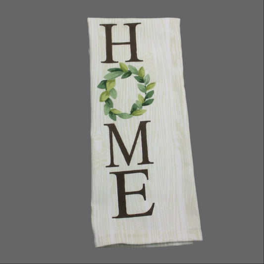 A rectangular white fabric towel with a wood-grain texture, featuring the word 'HOME' in dark brown letters, where the 'O' is replaced by a green leaf wreath. The towel is laid flat on a dark gray fabric background.