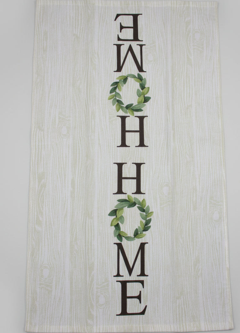 A rectangular white fabric tea towel with a light wood grain print, featuring the word 'HOME' spelled out in dark brown letters, with the 'O's replaced by green leaf wreaths. The towel is laid flat fully opened on a dark gray fabric surface.
