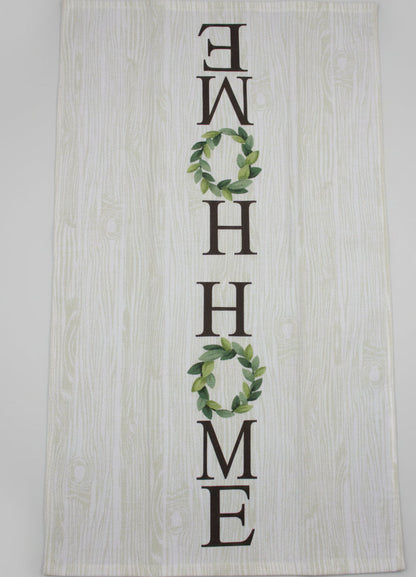 A rectangular white fabric tea towel with a light wood grain print, featuring the word 'HOME' spelled out in dark brown letters, with the 'O's replaced by green leaf wreaths. The towel is laid flat fully opened on a dark gray fabric surface.