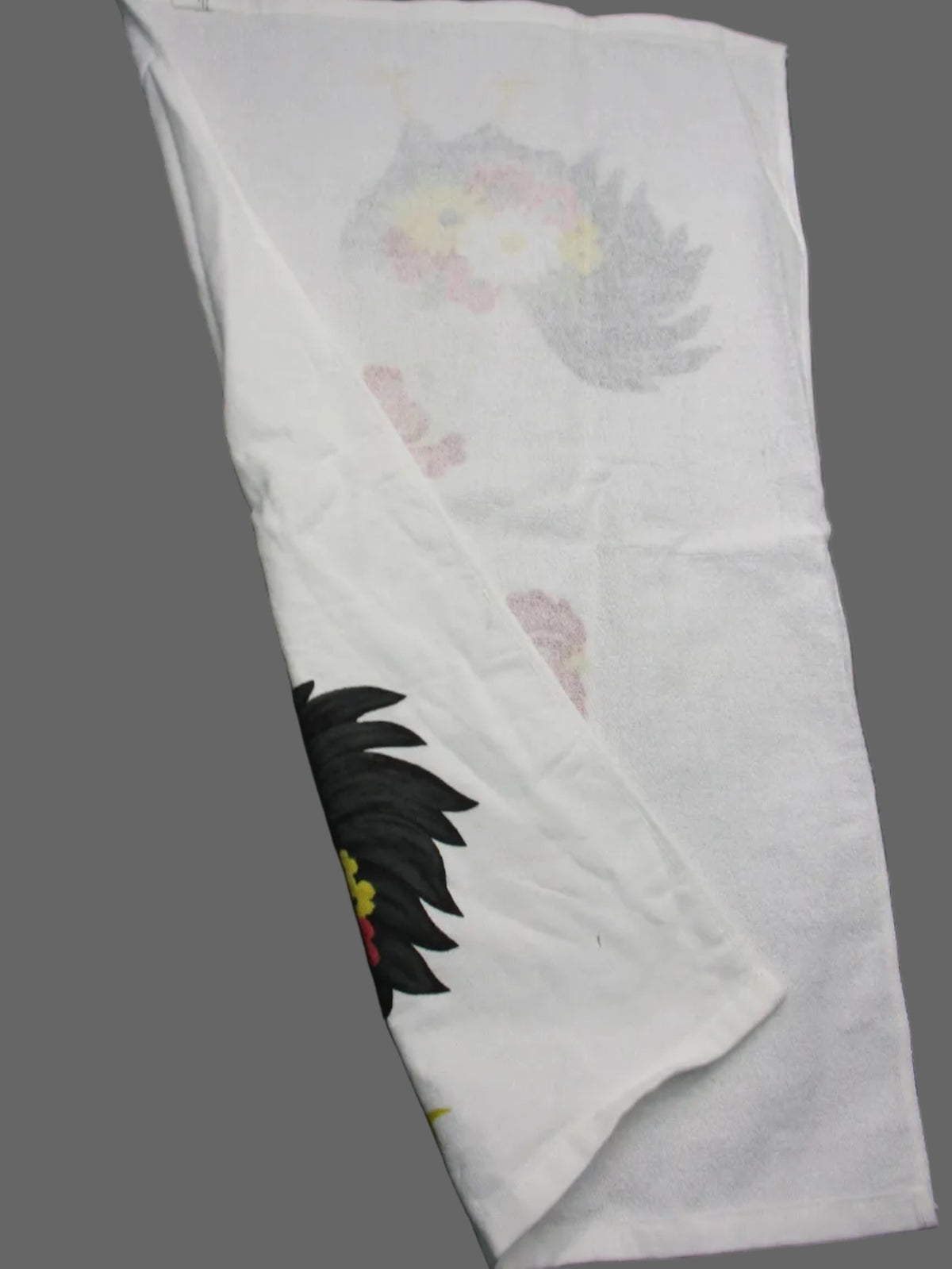Back side of towel, featuring a faded gray and pink floral pattern on one side and a black leaf or feather-like graphic with small yellow and red accents on the other, displayed on a dark gray background.