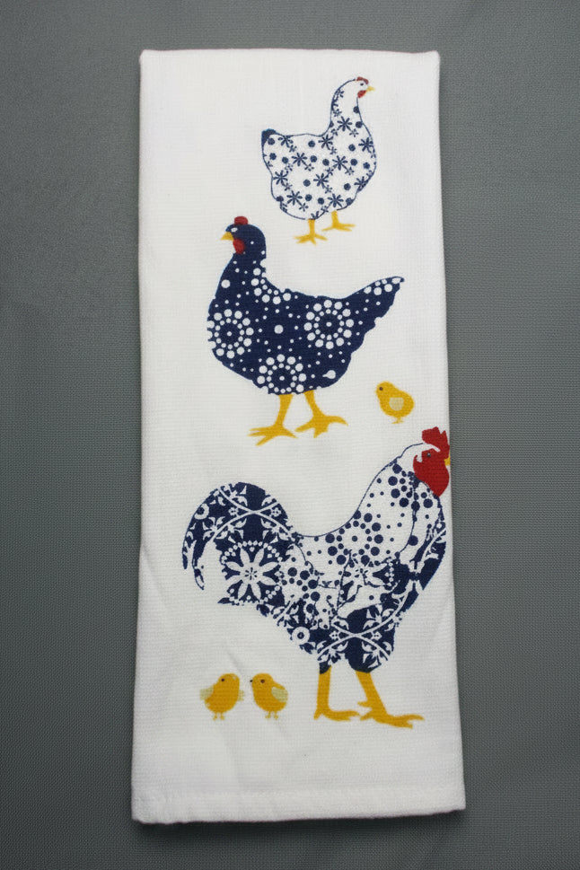 A folded white kitchen towel featuring a printed pattern of three chickens and two yellow chicks in navy blue and white floral designs, set against a dark gray background.