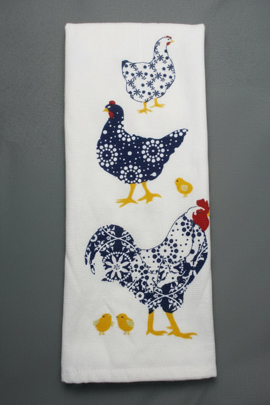 A folded white kitchen towel featuring a printed pattern of three chickens and two yellow chicks in navy blue and white floral designs, set against a dark gray background.