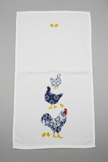 A white fabric towel featuring  chickens and chicks in navy, white, and yellow, laid opened flat on a gray surface.