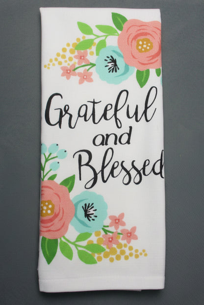 A folded white fabric item, likely a tea towel, printed with the words 'Grateful and Blessed' in black script, surrounded by a colorful floral design in pink, teal, yellow, and green, laid flat on a dark gray background.