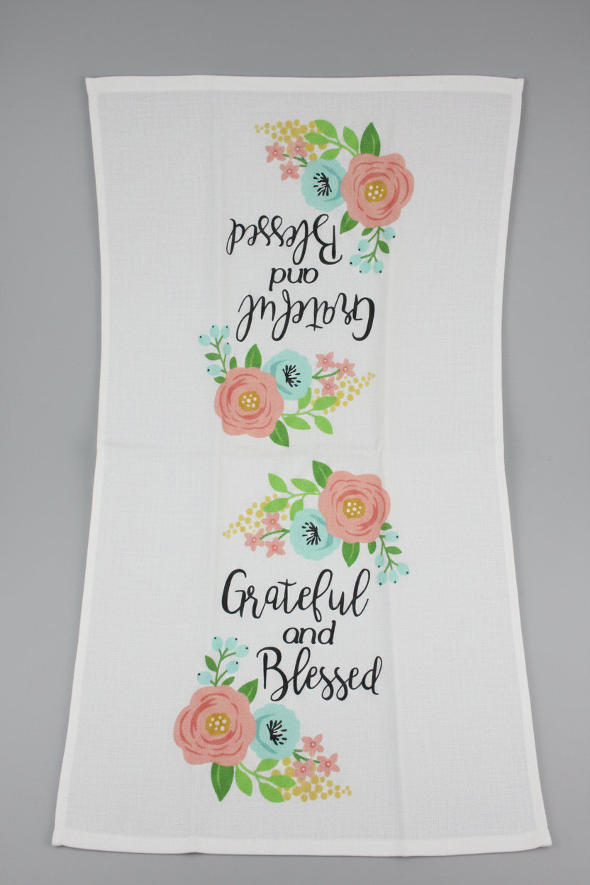 A white towel opened fully featuring the printed phrase 'Grateful and Blessed' in black script, surrounded by pink flowers, green leaves, and light blue and yellow floral accents.