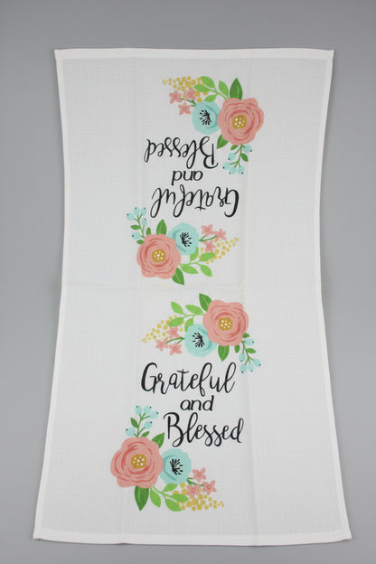 A white towel opened fully featuring the printed phrase 'Grateful and Blessed' in black script, surrounded by pink flowers, green leaves, and light blue and yellow floral accents.
