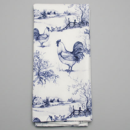 A folded white towel featuring a classic blue toile de Jouy pattern with roosters, trees, and rustic details, laid flat on a dark gray textured background.