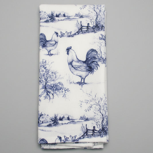 A folded white towel featuring a classic blue toile de Jouy pattern with roosters, trees, and rustic details, laid flat on a dark gray textured background.
