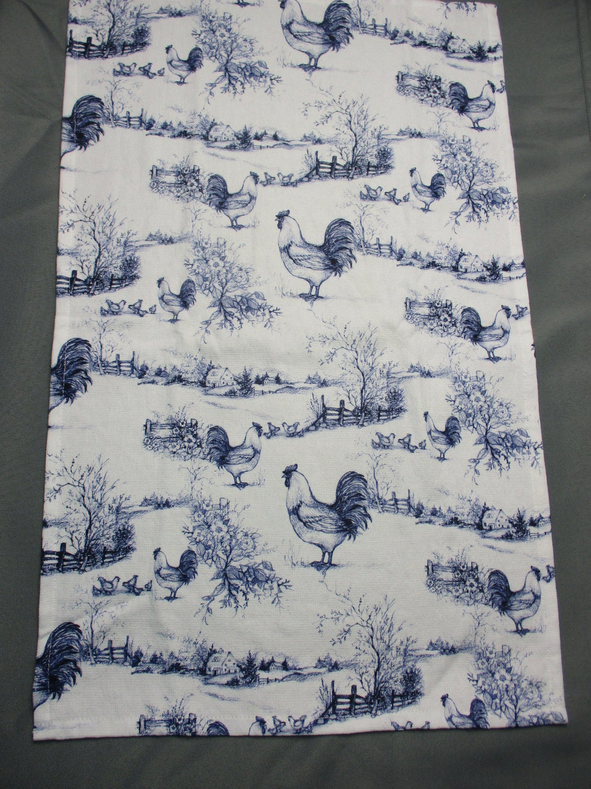 Towel fully opened featuring a repeating blue toile-style print of roosters, trees, and garden scenes, laid flat on a dark gray surface.
