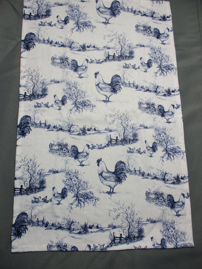 Towel fully opened featuring a repeating blue toile-style print of roosters, trees, and garden scenes, laid flat on a dark gray surface.