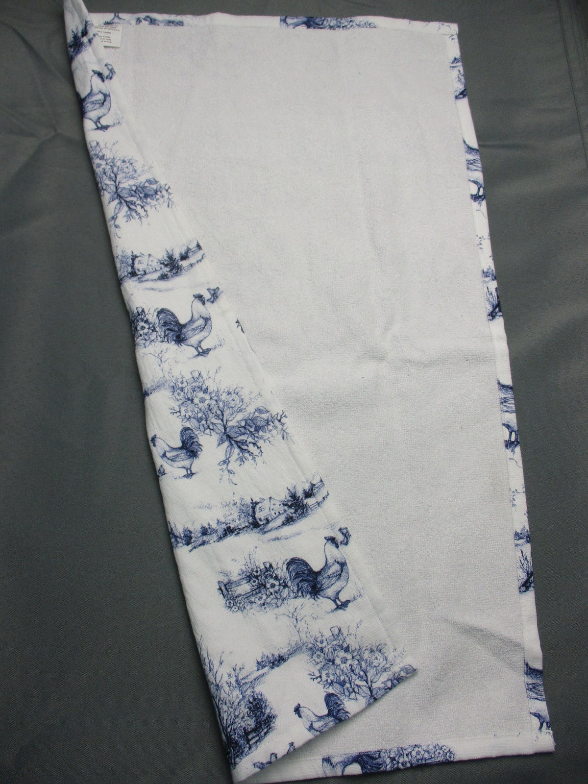 Back side of towel, featuring a classic blue toile de Jouy pattern of animals and trees, laid flat on a dark gray surface.
