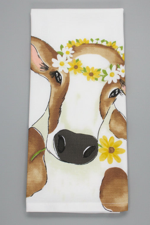 A folded white kitchen towel featuring a watercolor-style illustration of a cow's face adorned with a yellow and white flower crown, set against a dark gray background.