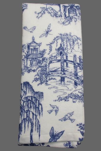 A folded white towel, featuring a classic blue toile de Jouy pattern with pagodas, butterflies, and foliage, displayed on a dark gray background.
