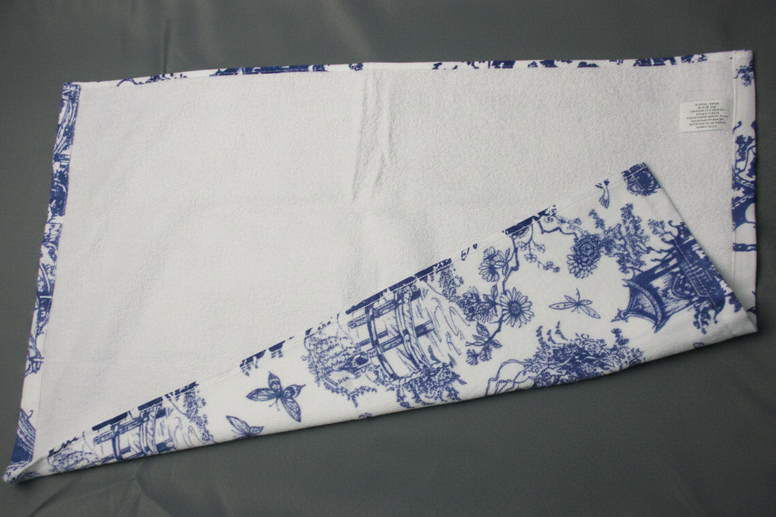 Back side of towel with a blue toile de Jouy pattern, folded diagonally on a dark gray fabric background.