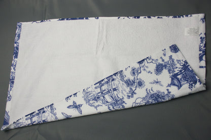 Back side of towel with a blue toile de Jouy pattern, folded diagonally on a dark gray fabric background.