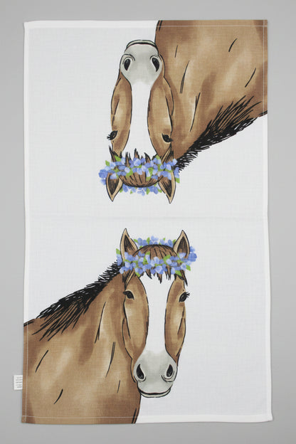 A white  towel fully opened featuring a printed design of two brown horses facing each other, each wearing a blue flower crown, laid flat on a dark gray fabric surface.