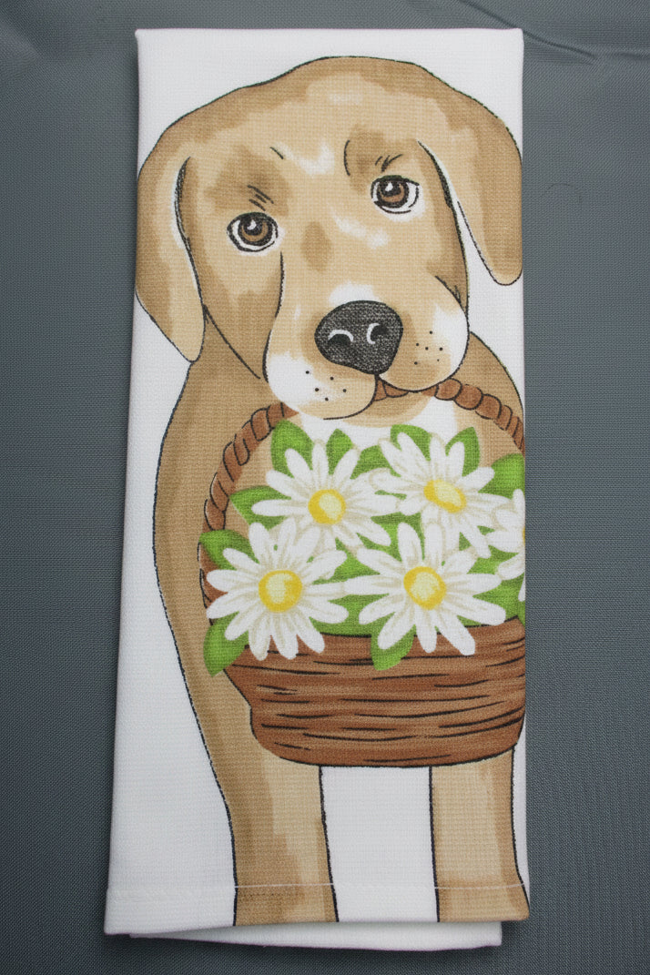 A folded white kitchen towel featuring a printed illustration of a tan dog holding a brown basket of white daisies with green leaves, laid flat on a dark gray background.
