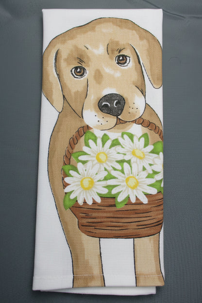 A folded white kitchen towel featuring a printed illustration of a tan dog holding a brown basket of white daisies with green leaves, laid flat on a dark gray background.