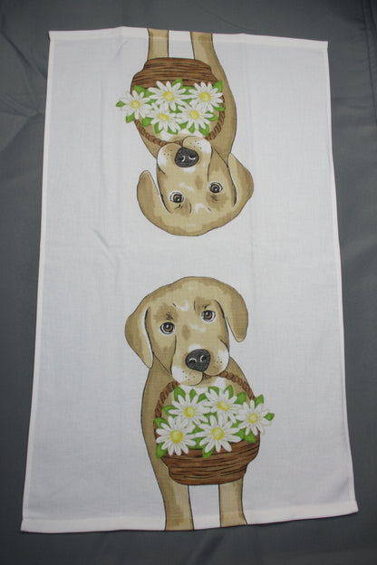 A white towel laid fully opened flat on a dark gray surface, printed with two cartoon dogs facing each other and each holding a basket of white daisies with green leaves.