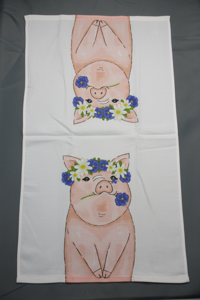 A white rectangular towel fully opened on a dark gray surface, printed with two cartoon pigs facing inward and wearing blue, white, and yellow flower crowns.