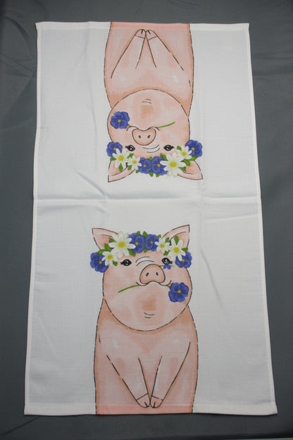 A white rectangular towel fully opened on a dark gray surface, printed with two cartoon pigs facing inward and wearing blue, white, and yellow flower crowns.