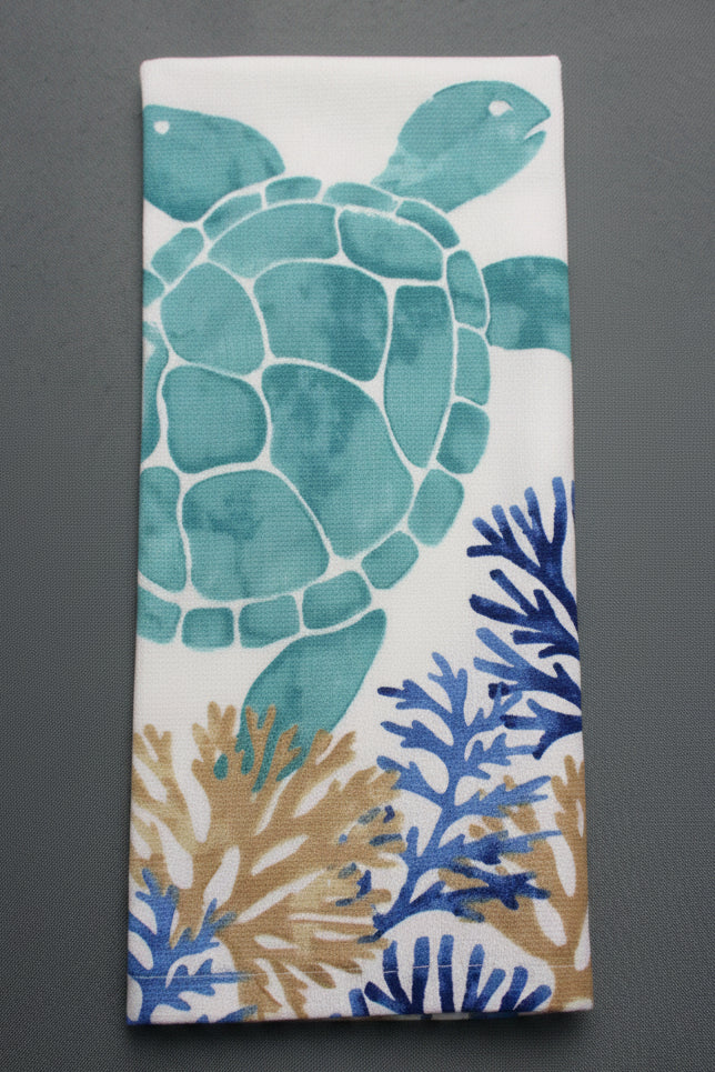 A folded white towel, featuring a printed design of a teal-green sea turtle and blue and tan coral motifs, displayed on a dark gray background.