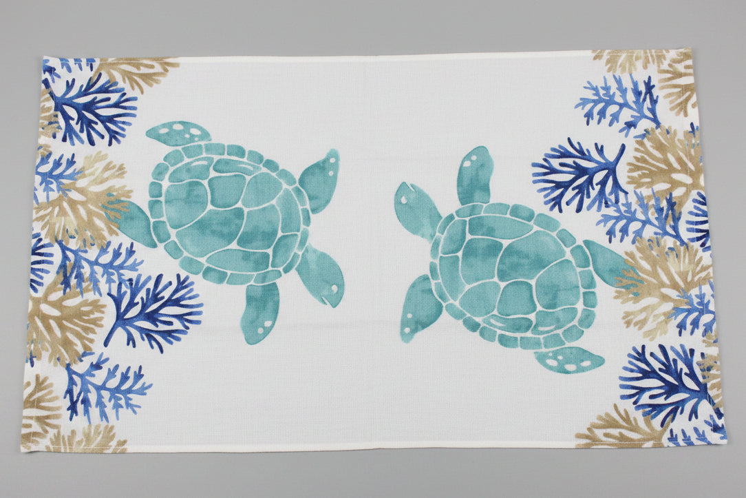 A rectangular white towel fully opened featuring a watercolor-style print of two teal sea turtles and blue and tan coral motifs along the borders, laid flat on a dark gray surface.