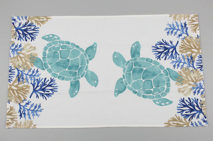 A rectangular white towel fully opened featuring a watercolor-style print of two teal sea turtles and blue and tan coral motifs along the borders, laid flat on a dark gray surface.