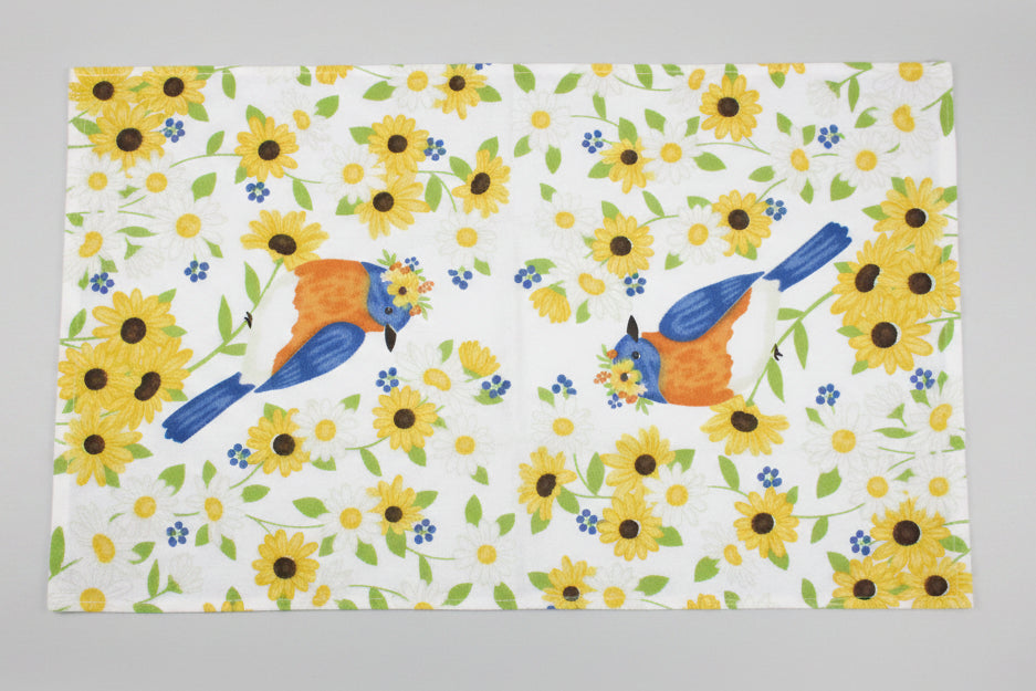Fully opened towel featuring a white background printed with yellow sunflowers, green leaves, and two blue-and-orange birds, laid flat on a gray surface.