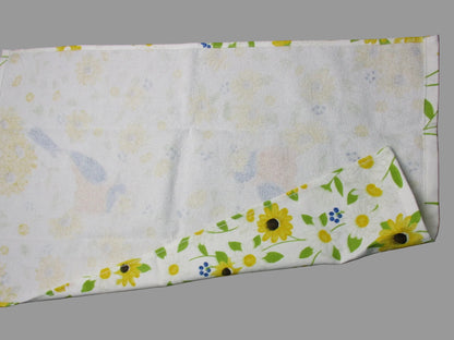 Back side of towel, featuring a floral print with yellow sunflowers, green leaves, and small blue accents. The fabric is laid flat on a dark gray surface with a metal ruler visible on the right edge.