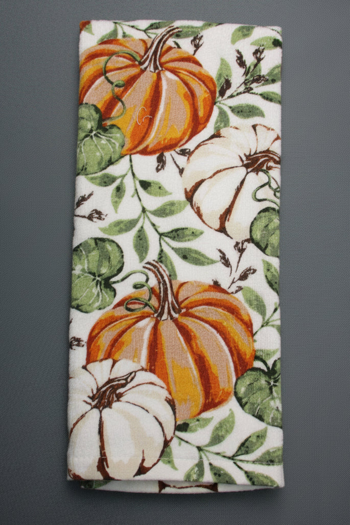 A towel, featuring a white background with a printed autumn pattern of orange and white pumpkins and green leaves, laid flat on a dark gray surface.