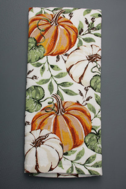 A towel, featuring a white background with a printed autumn pattern of orange and white pumpkins and green leaves, laid flat on a dark gray surface.